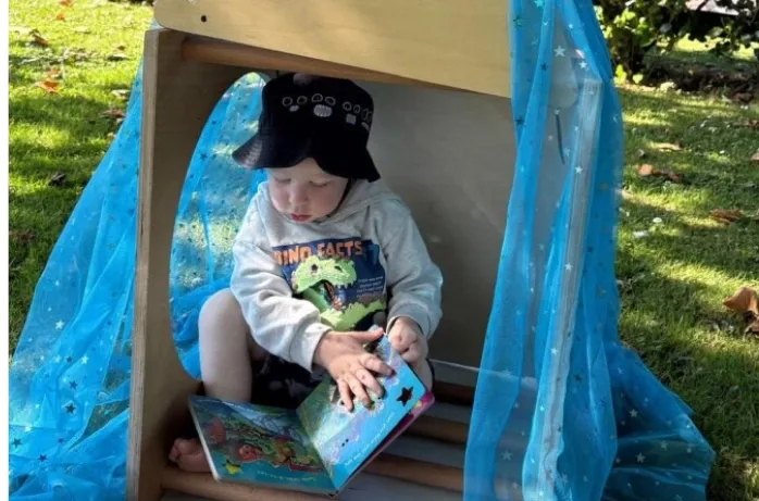 A toddler in a sunhat reads a picture book inside a small play hut draped with blue fabric on the lawn.