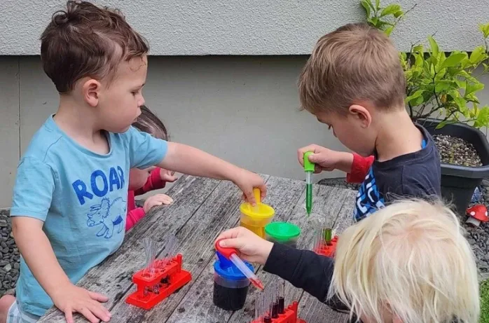 Three young children use pipettes and coloured water at an outdoor table.