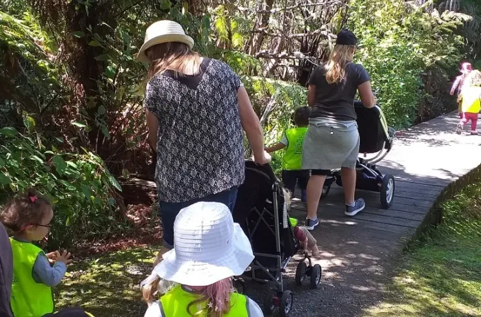 Educators with prams and young children in hi-vis vests walk along a bush boardwalk.