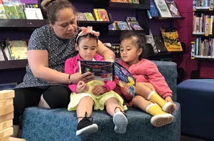 An educator reads a picture book with two young girls on a couch in a library.