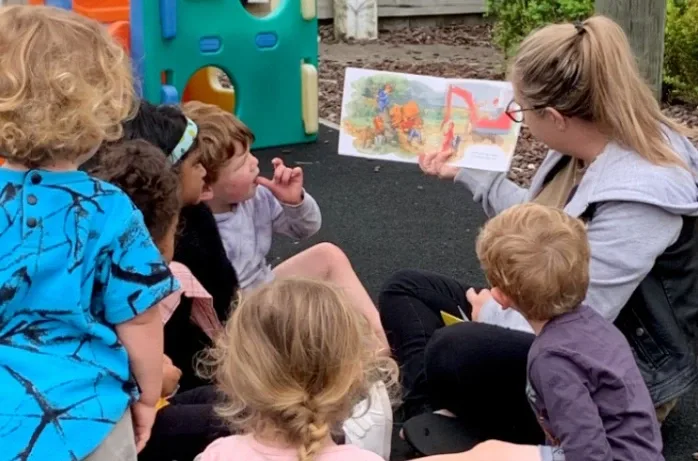 An educator reads a picture book to a group of children sitting outdoors.