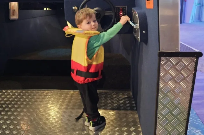 A young boy in a life jacket turns a dial beside a ship's wheel display.