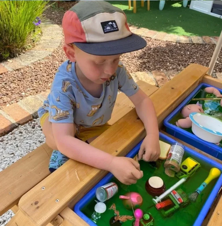 A boy in a cap plays with jars and toys in a green water tray outdoors.