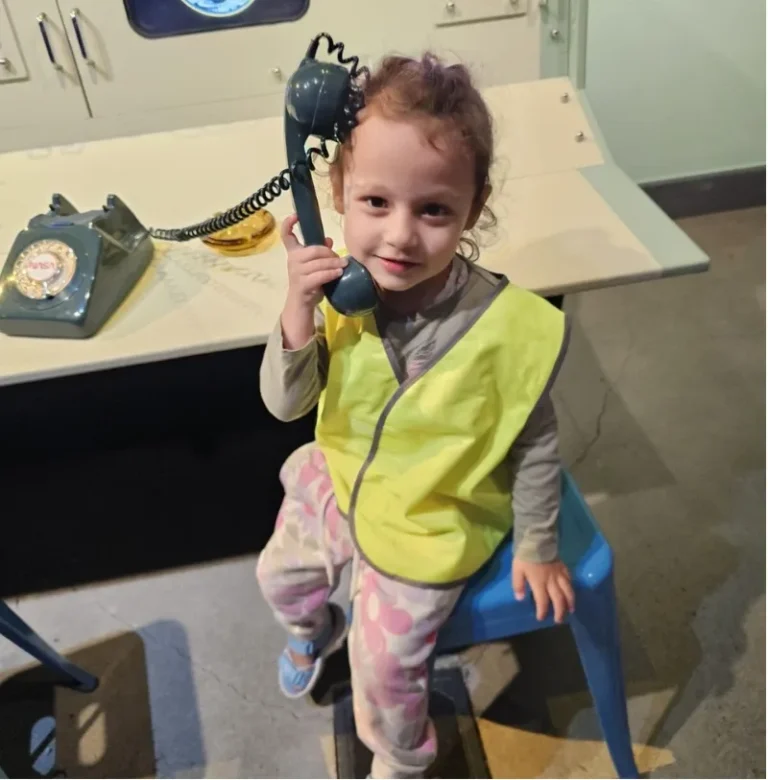 A girl in a hi-vis vest holds an old-fashioned telephone to her ear.