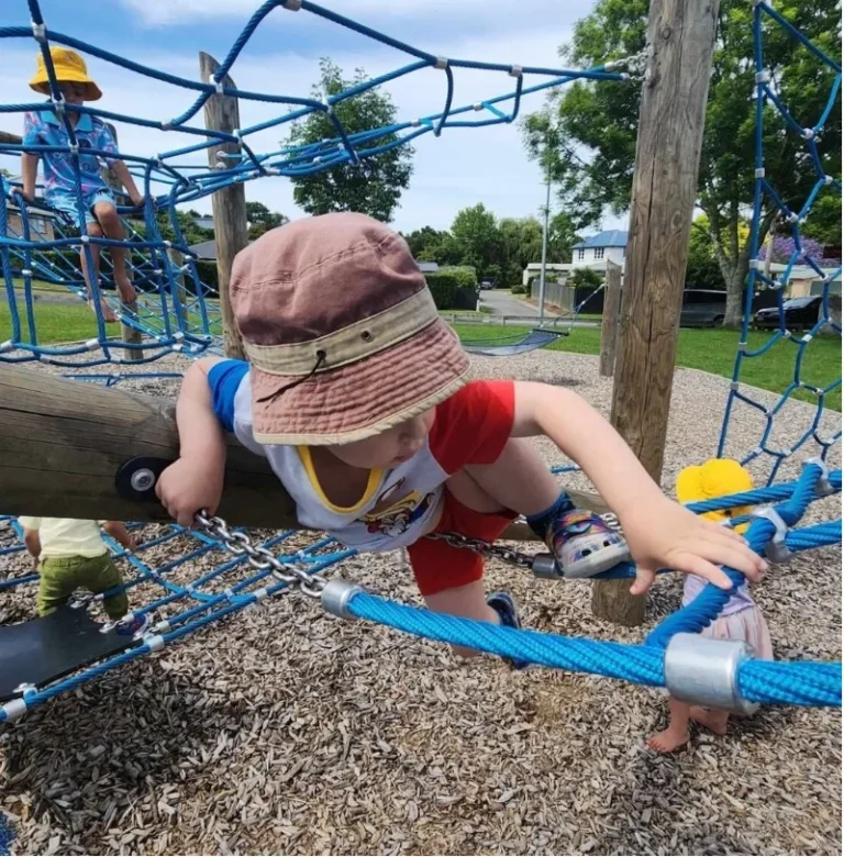 A toddler in a sunhat climbs across a rope net in a playground.
