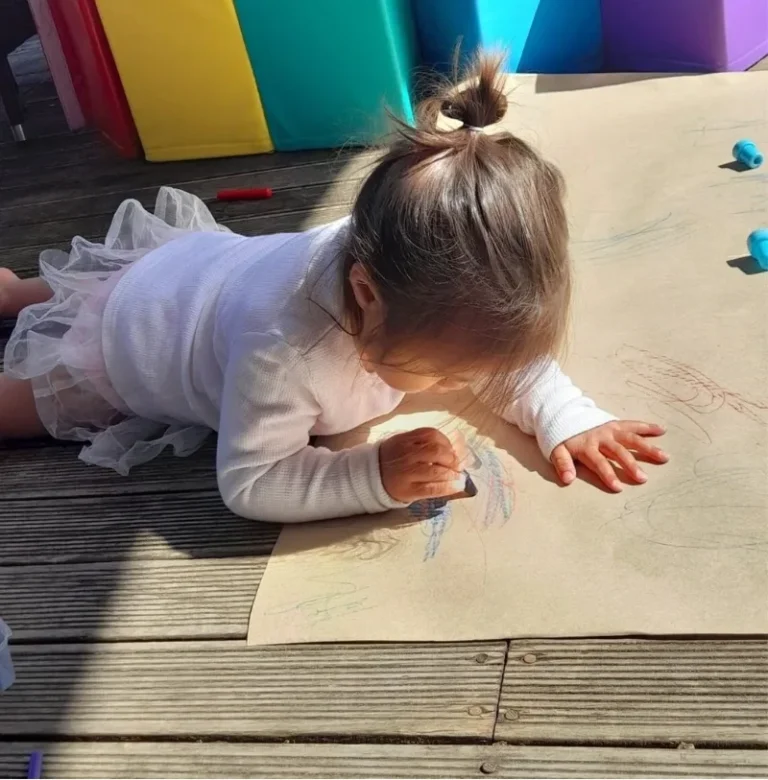 A toddler lies on a deck drawing with crayons on a large sheet of paper.