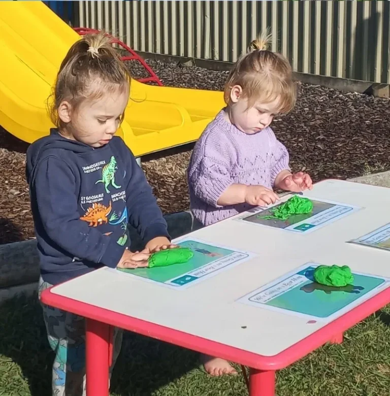 Two toddlers roll green play dough on mats at an outdoor table.