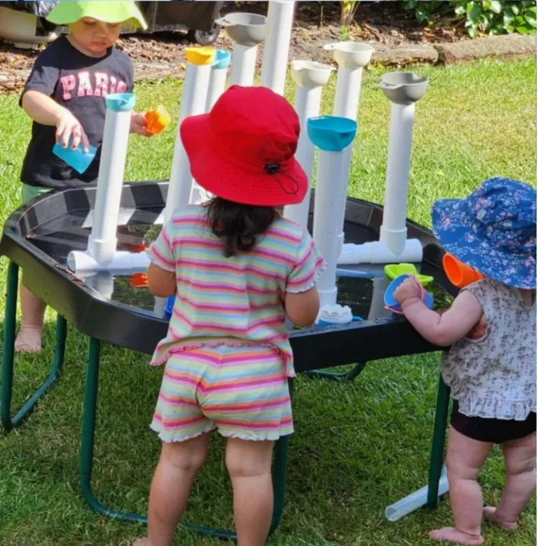 Toddlers in sunhats pour water through pipes at an outdoor water play station.