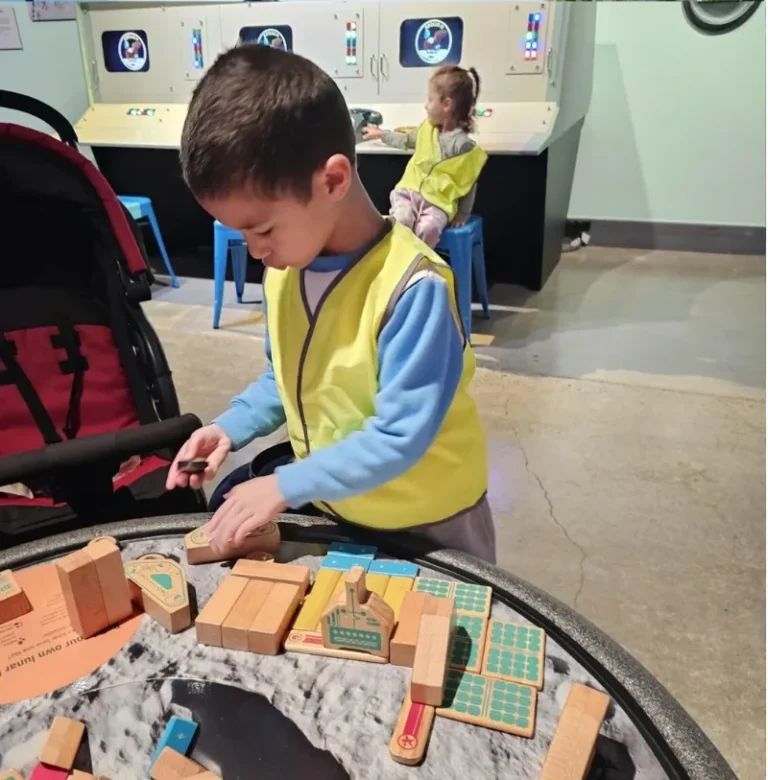 A boy in a hi-vis vest builds with wooden blocks in a tray of sand.