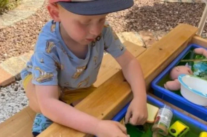 A boy in a cap plays with toys in a water tray outdoors.