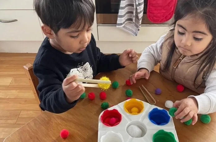 Two young children use tongs to sort coloured pom-poms into a matching colour tray.