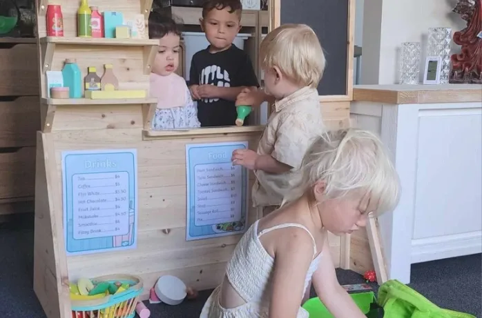 Young children play shop at a wooden play-shop stand with price lists.
