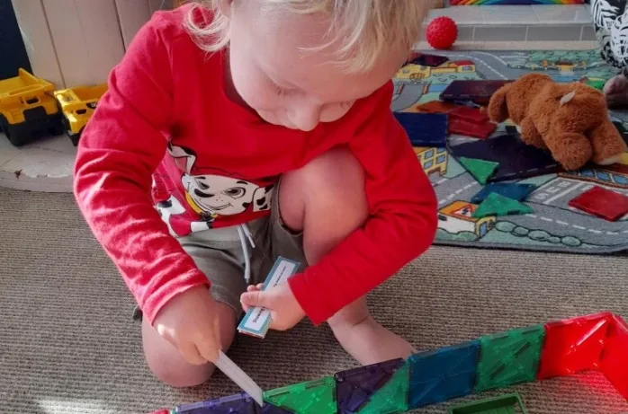 A toddler builds a line of coloured magnetic tiles on the floor.