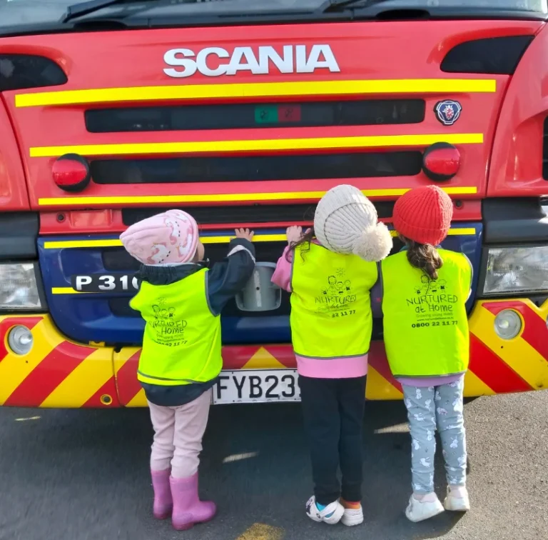 Three young children in hi-vis vests stand at the front of a fire engine.