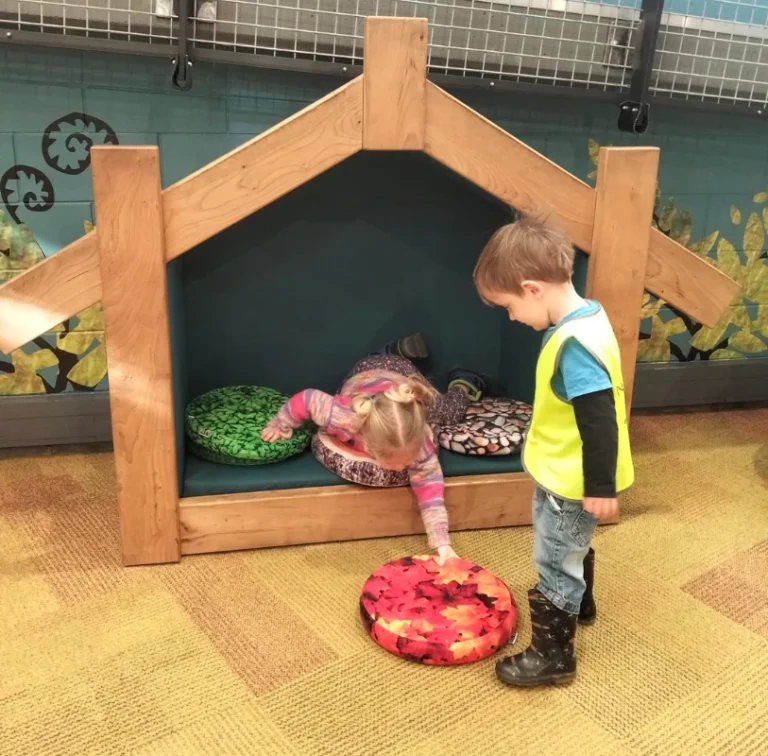 Two young children play with cushions in a wooden wharenui-style play house.