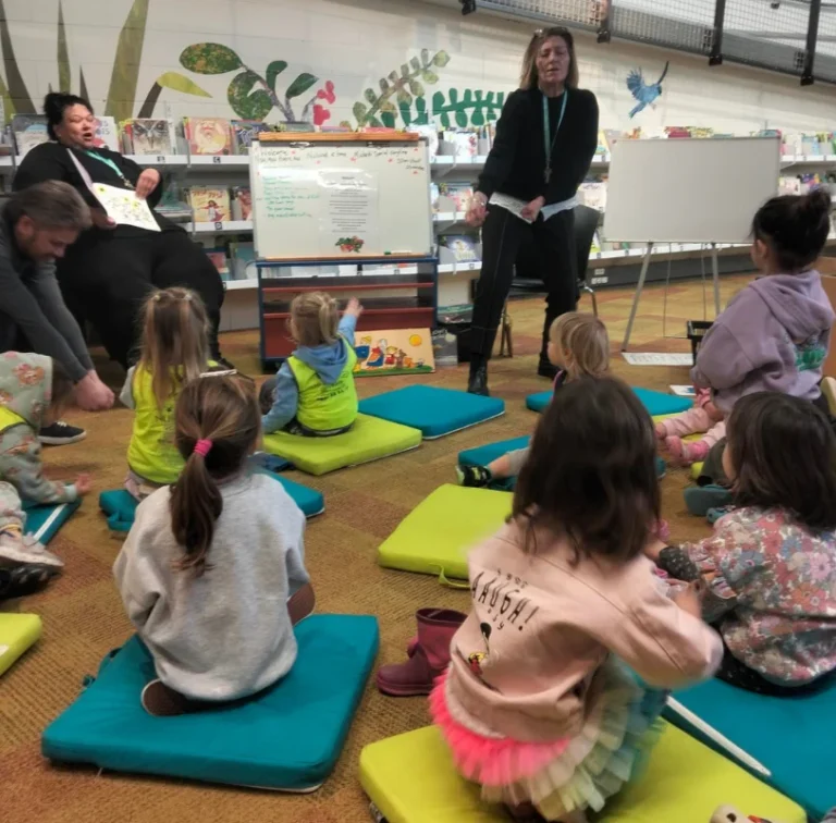 Children sit on cushions for a library session while adults look on.