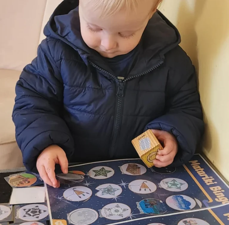 A toddler plays a Matariki bingo game with a wooden block.