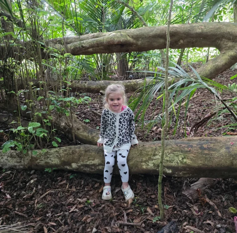 A smiling girl sits on a fallen tree branch in the bush.