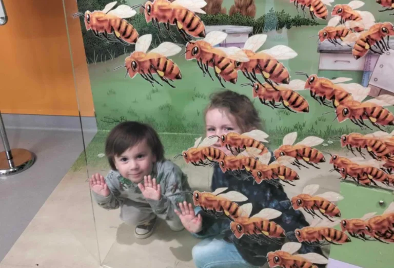A toddler and an adult press their hands against a glass display of giant bees.