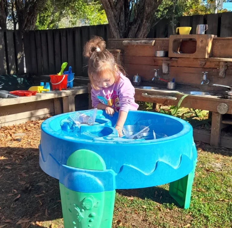 A toddler plays at a blue water table beside an outdoor mud kitchen.