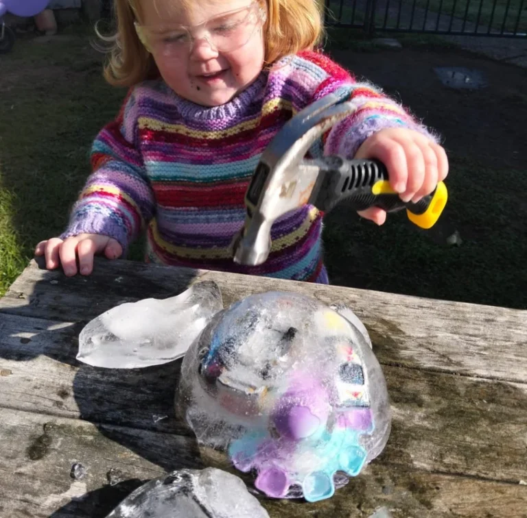 A toddler in safety glasses uses a hammer to break ice with toys frozen inside.