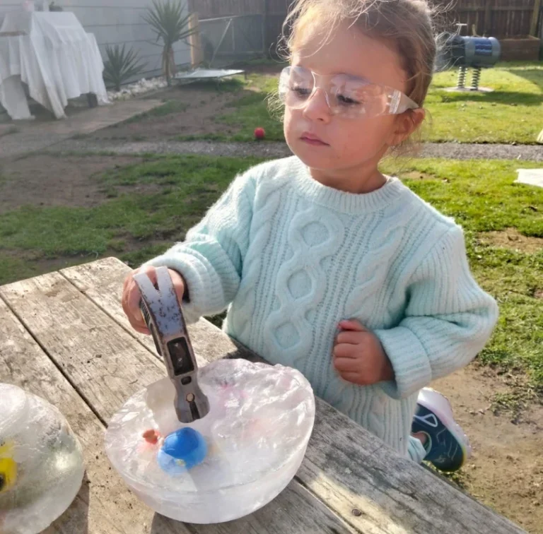 A toddler in safety glasses taps a bowl of ice with a hammer.