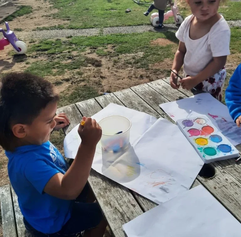 Young children paint with watercolours at an outdoor table.