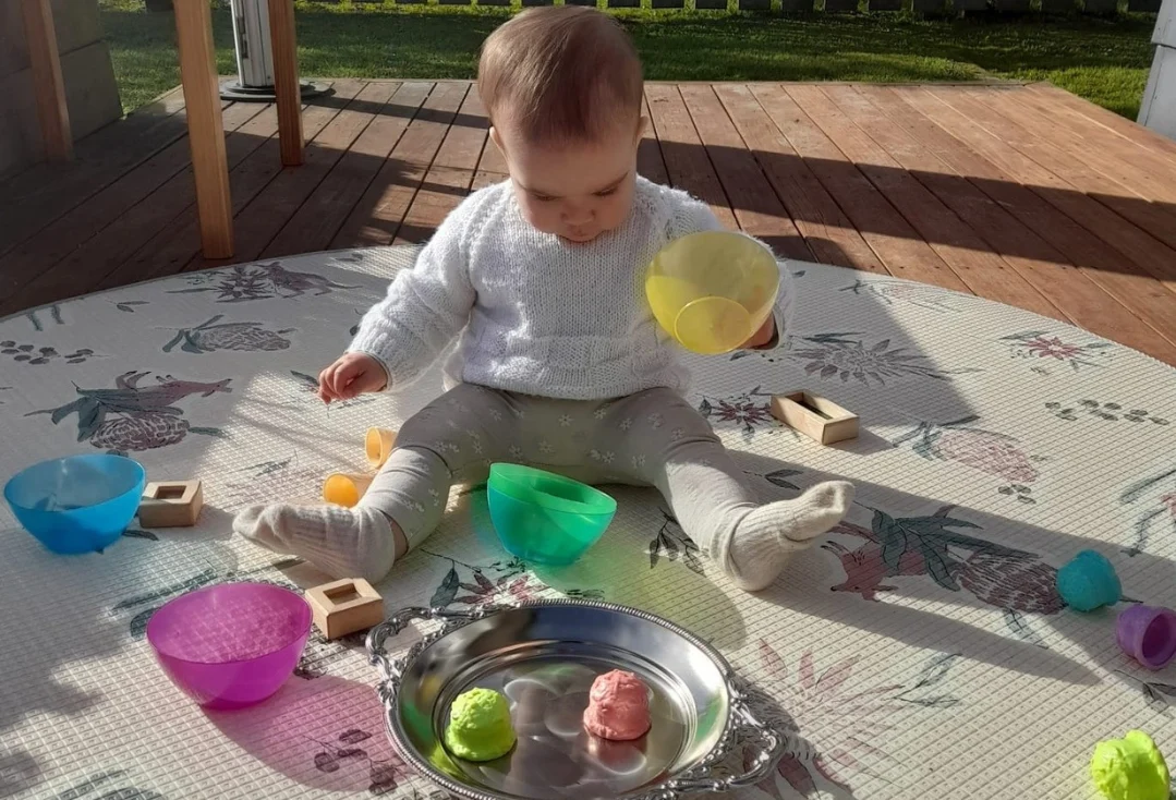 A baby sits on a play mat on a sunny deck, exploring coloured bowls, wooden blocks and a metal tray.