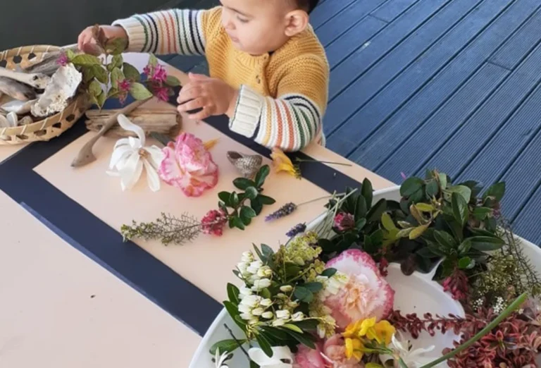 A baby explores flowers, leaves and shells laid out on a table on a deck.