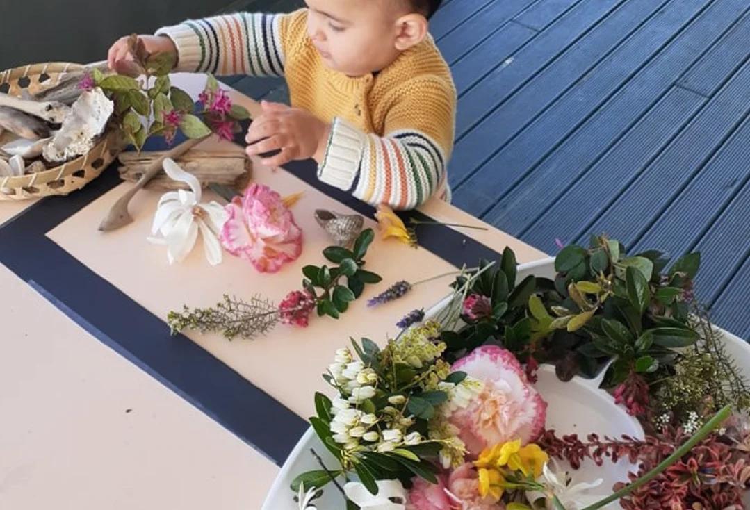 A baby explores flowers, leaves and shells laid out on a table on a deck.