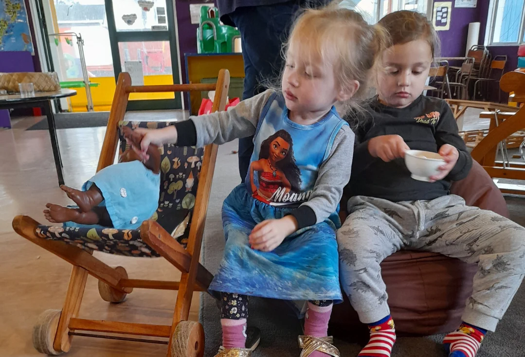 Two young children play at caring for a baby doll in a wooden pram and drinking from a toy teacup.