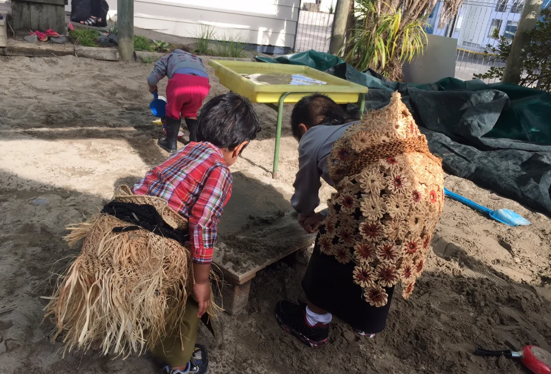 Children in dress-up costumes, including grass skirts and a woven cape, playing in a sandpit.