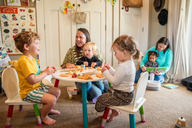 Children play with play dough at a small table while two educators and a baby sit alongside.