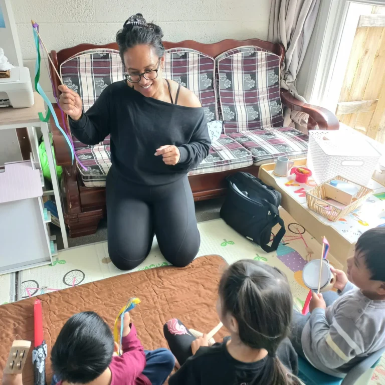 An educator kneels and waves ribbon sticks while young children play instruments on a mat.