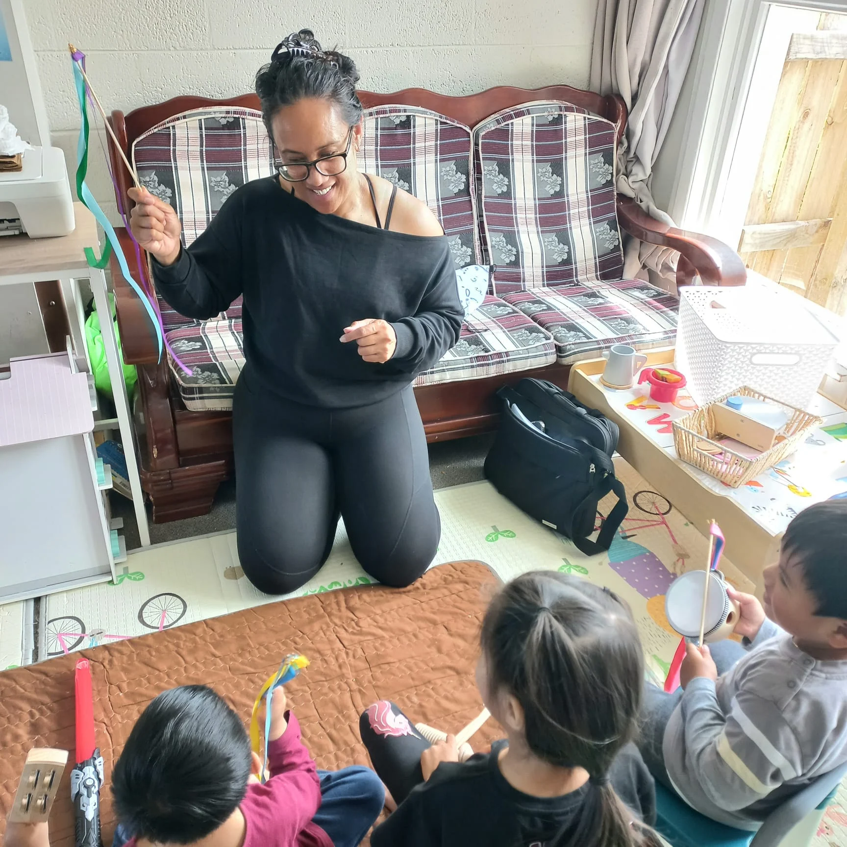 An educator kneels and waves ribbon sticks while young children play instruments on a mat.