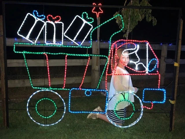 A young child stands inside a lit-up Christmas light display of a tractor carrying presents at night.
