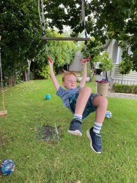A boy hangs from a rope swing under a tree in a garden.
