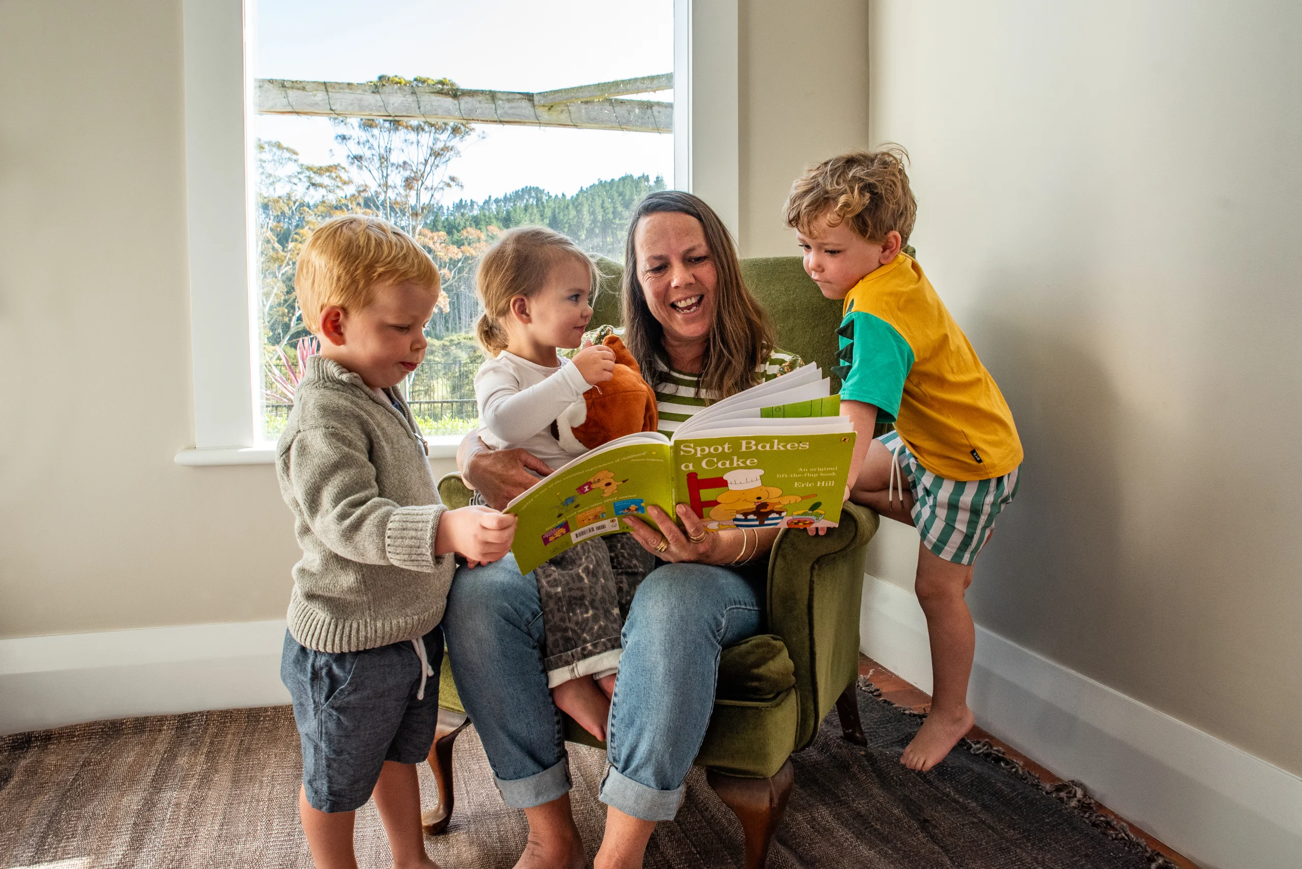 An educator in an armchair reads a picture book with three young children gathered around her.