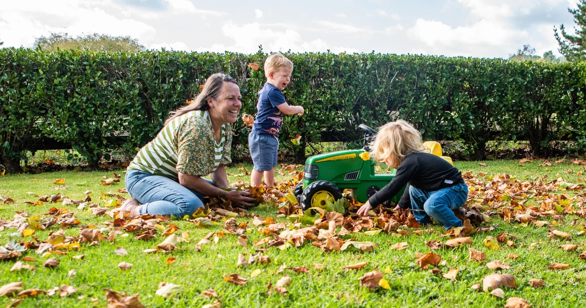An educator plays in autumn leaves on a lawn with two toddlers and a toy tractor.