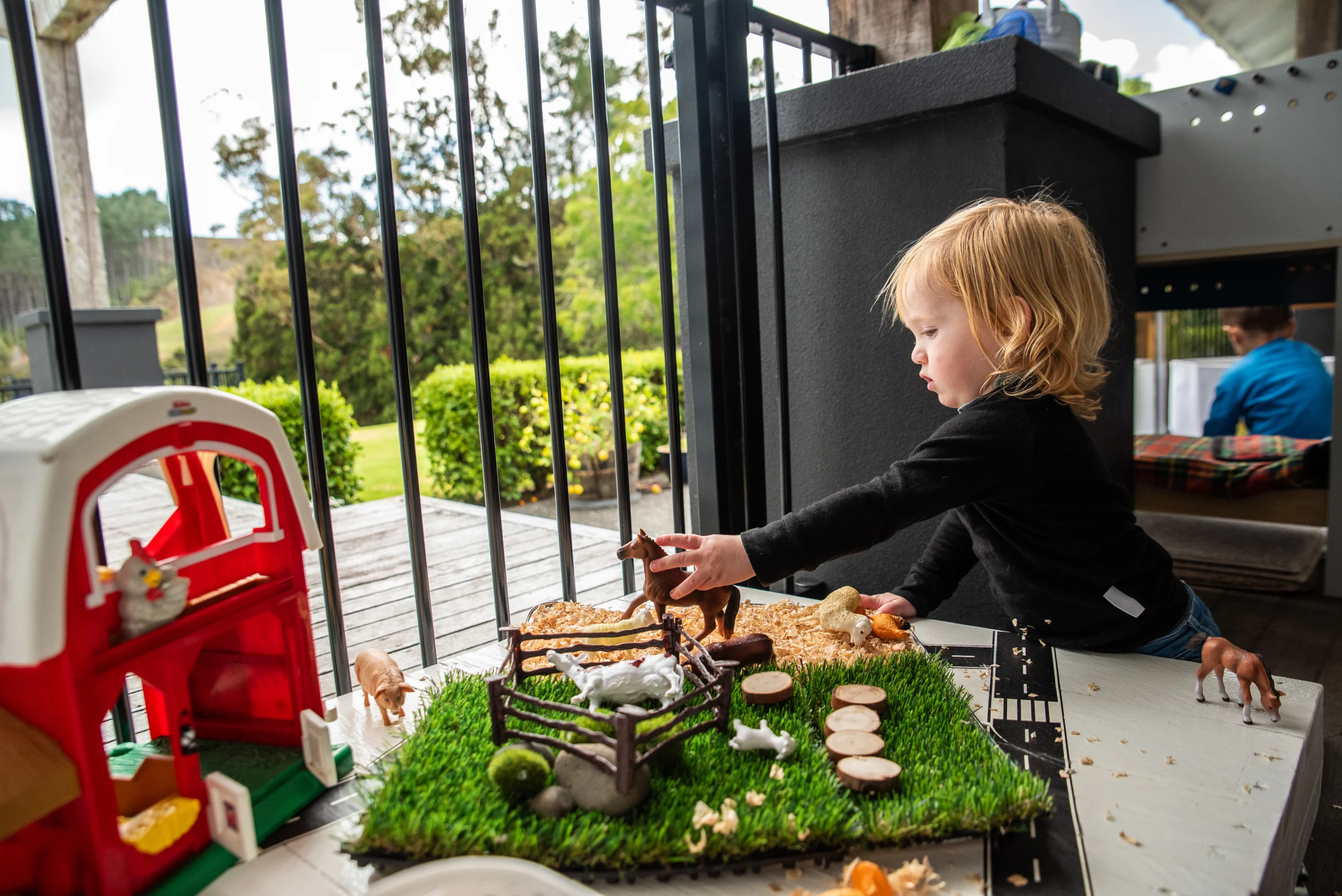 A toddler arranges toy farm animals on a small grass play mat beside a toy barn on a deck.