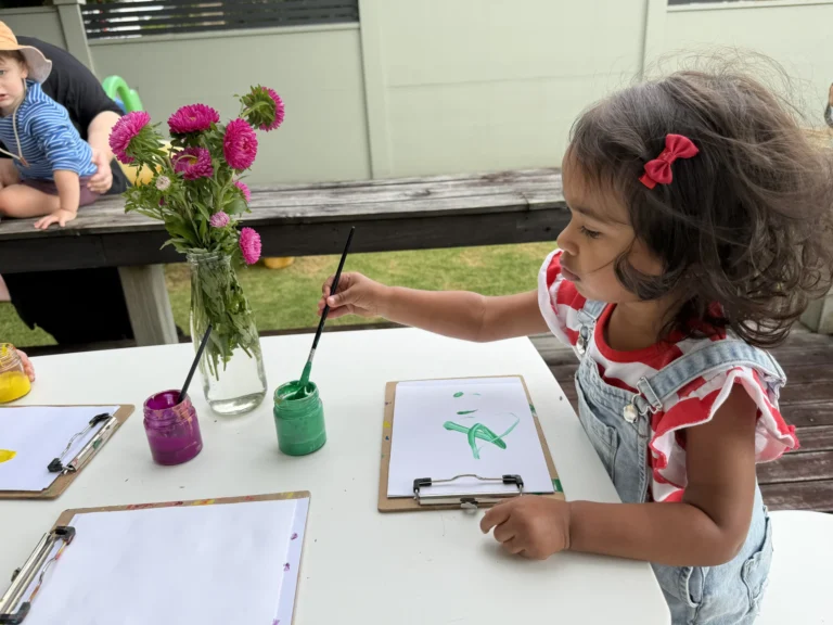 A young girl paints at an outdoor table set with paint pots, clipboards and a vase of flowers.