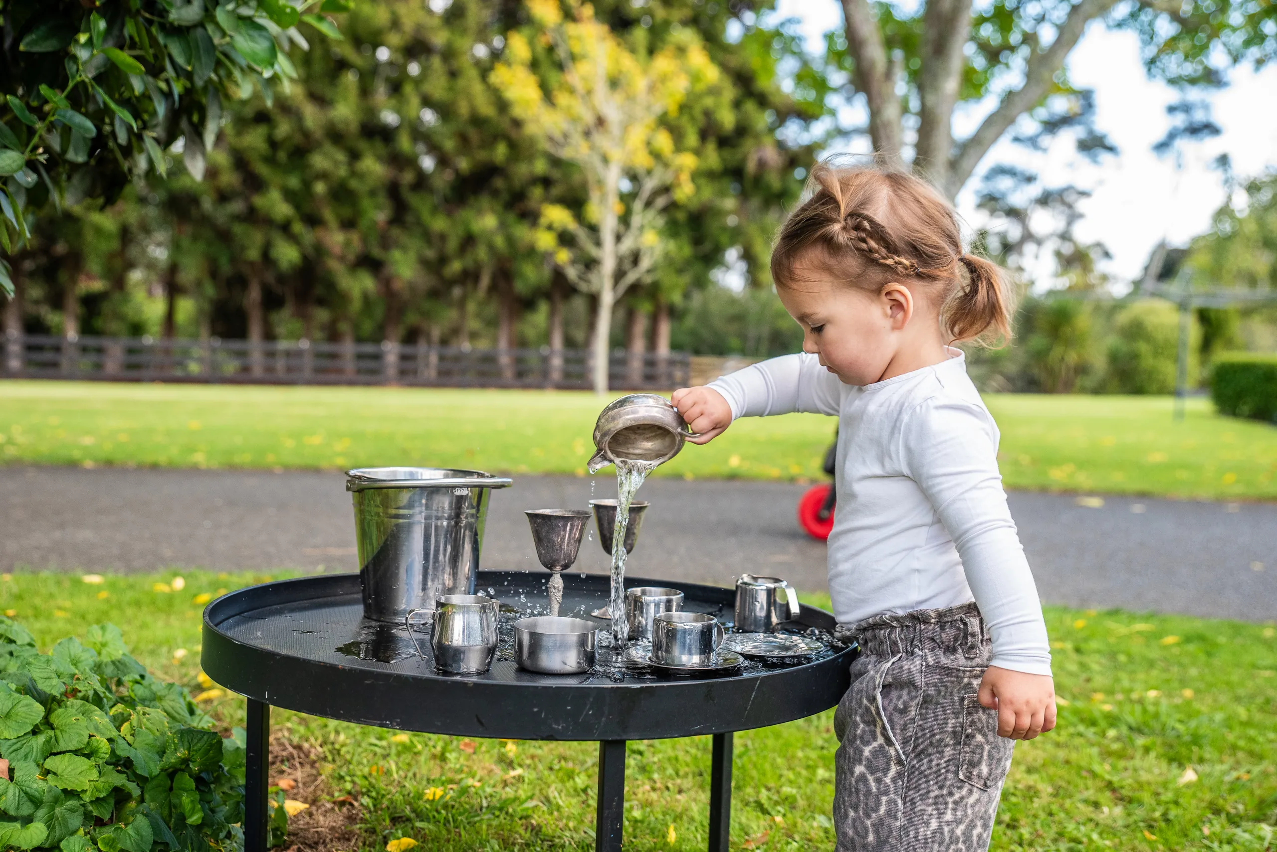 A toddler pouring water between metal cups at an outdoor play table