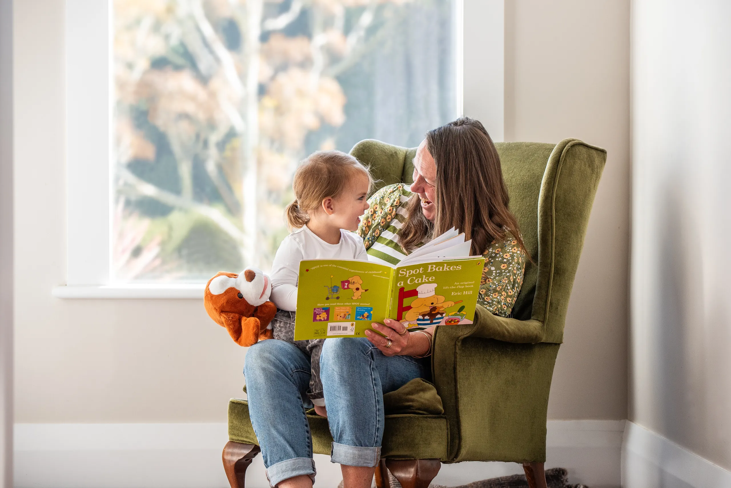 An Educator reading a picture book to a toddler in an armchair by a window