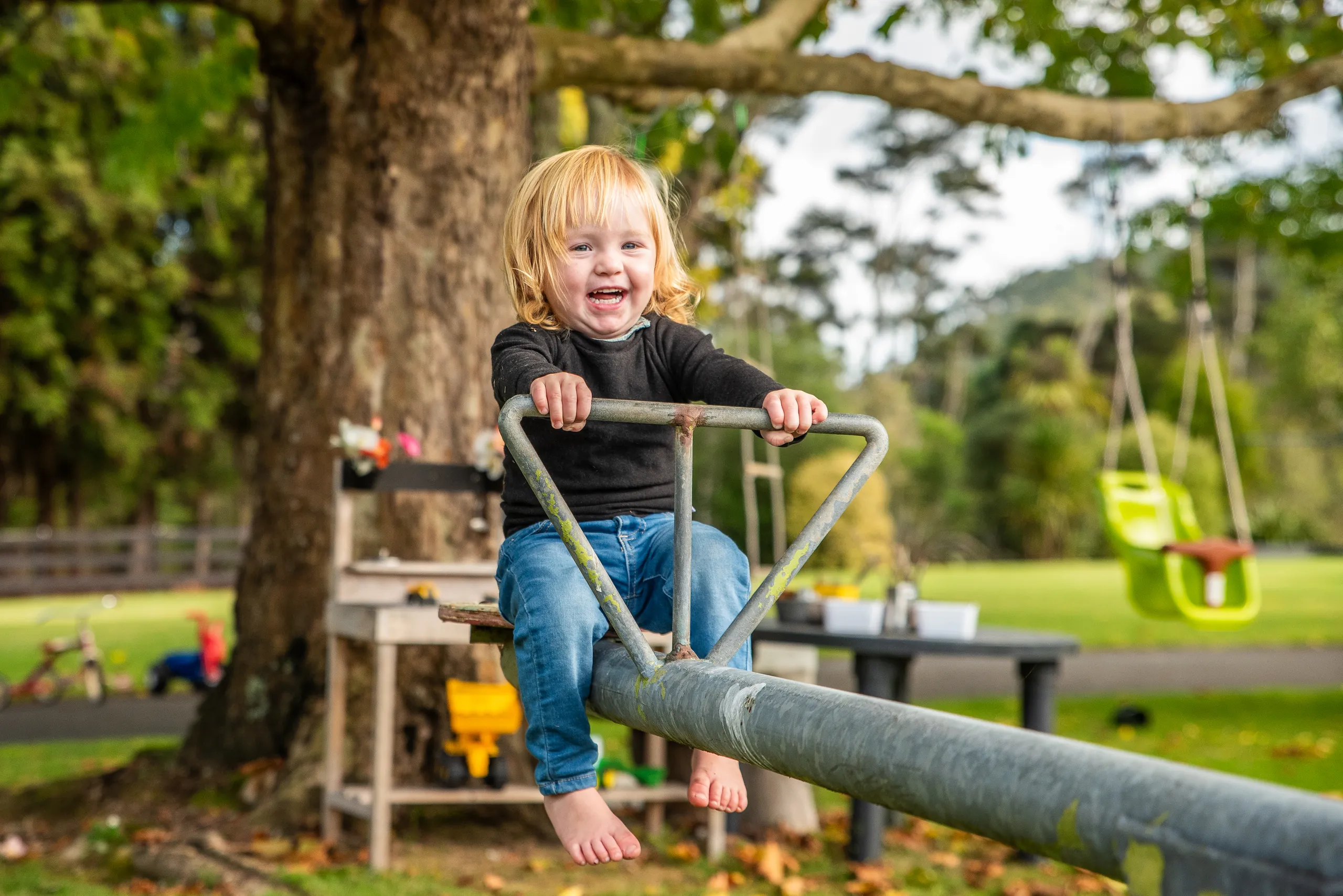 A smiling toddler riding a seesaw beneath a large tree