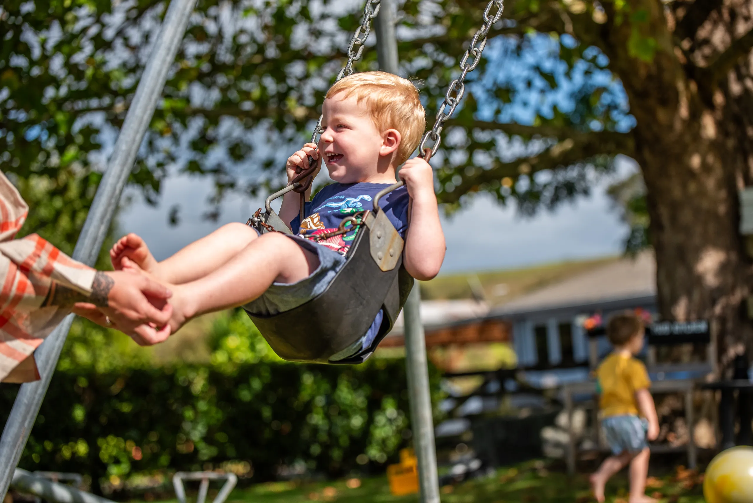 A laughing toddler on a swing being pushed, with an older child playing nearby