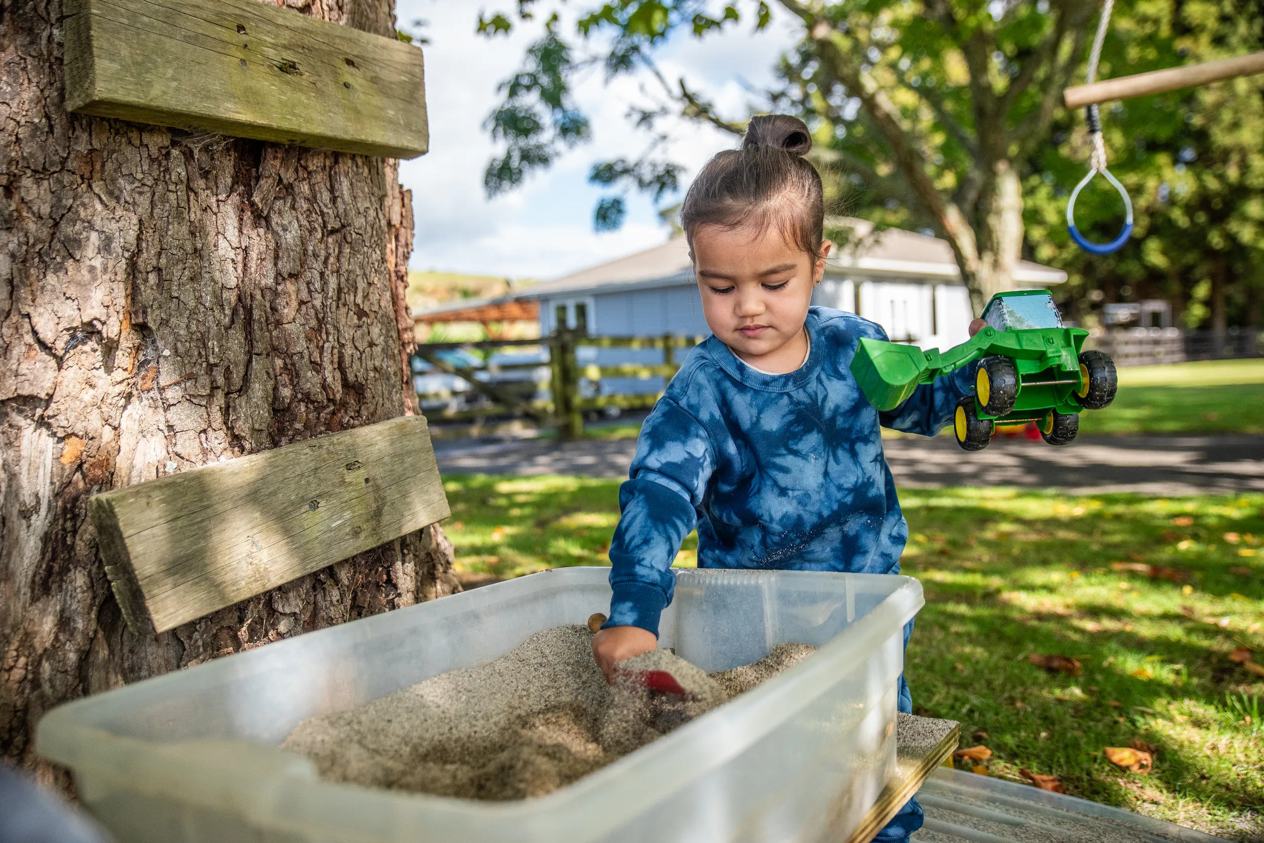 A toddler playing in a sandpit tub beside a tree in the garden