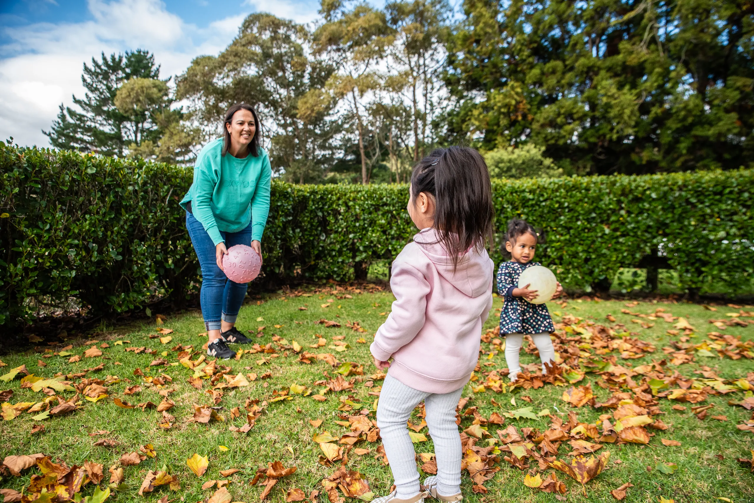 An Educator playing ball with two toddlers on a lawn covered in autumn leaves