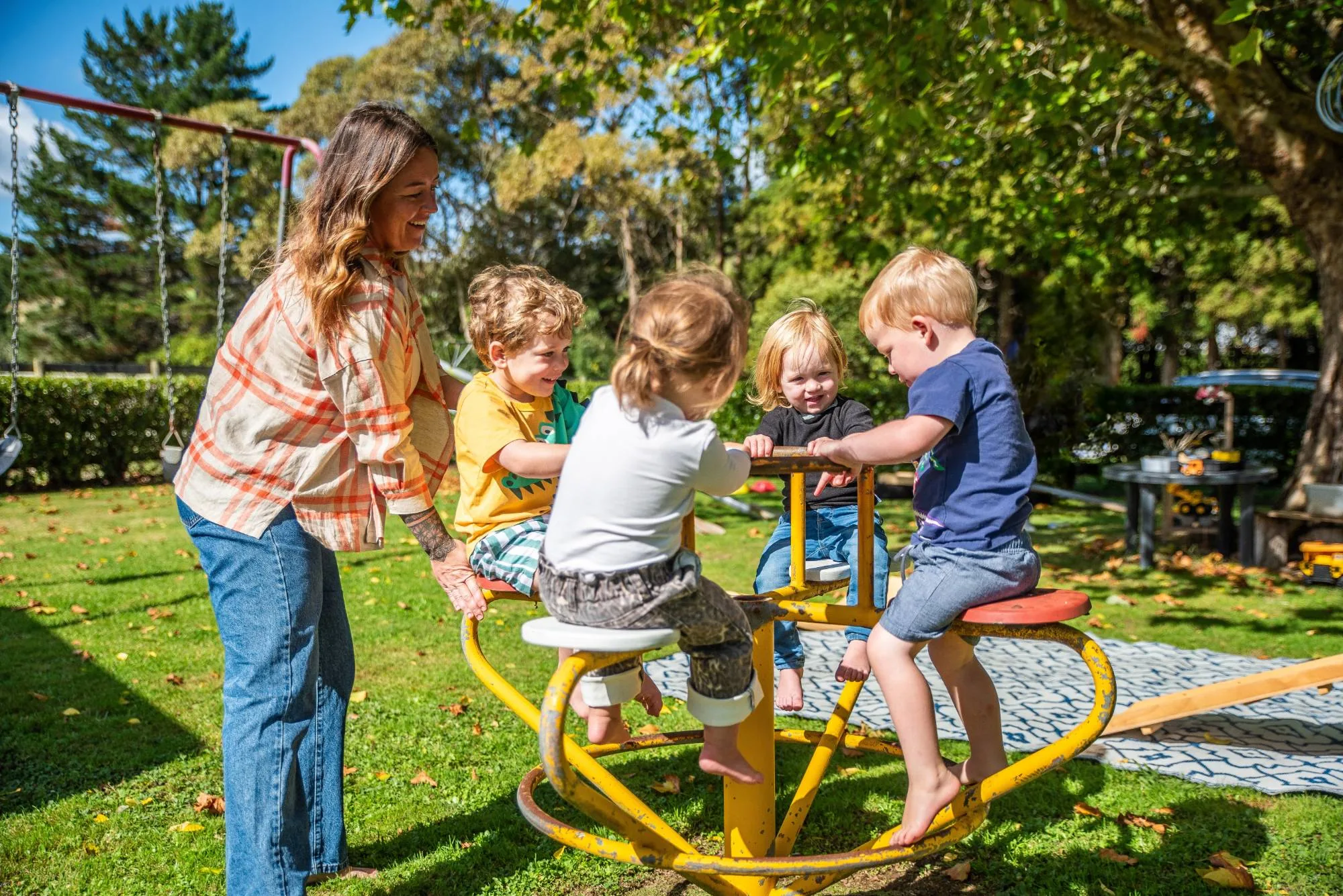 An Educator helping four toddlers on a yellow roundabout in a sunny garden